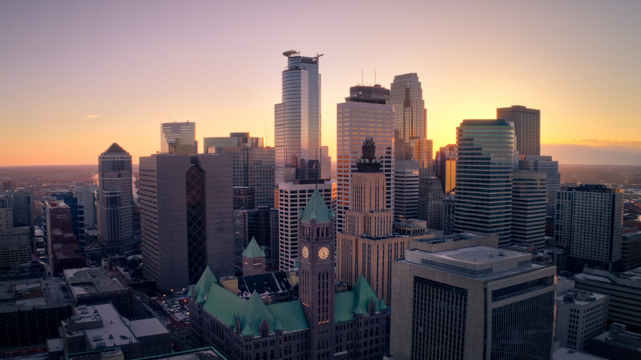 Minneapolis skyline at sunset, with tall buildings against a colorful orange and yellow sky.