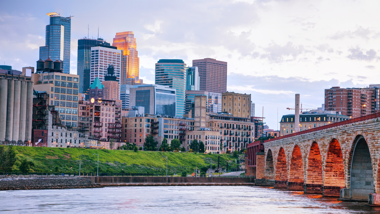 Minneapolis skyline with stone arch bridge and river under cloudy sky.