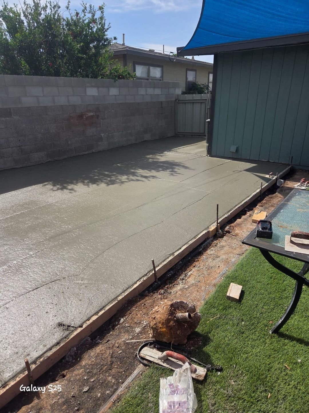 Freshly poured concrete driveway in a backyard, bordered by wood, with a blue shade and greenery.