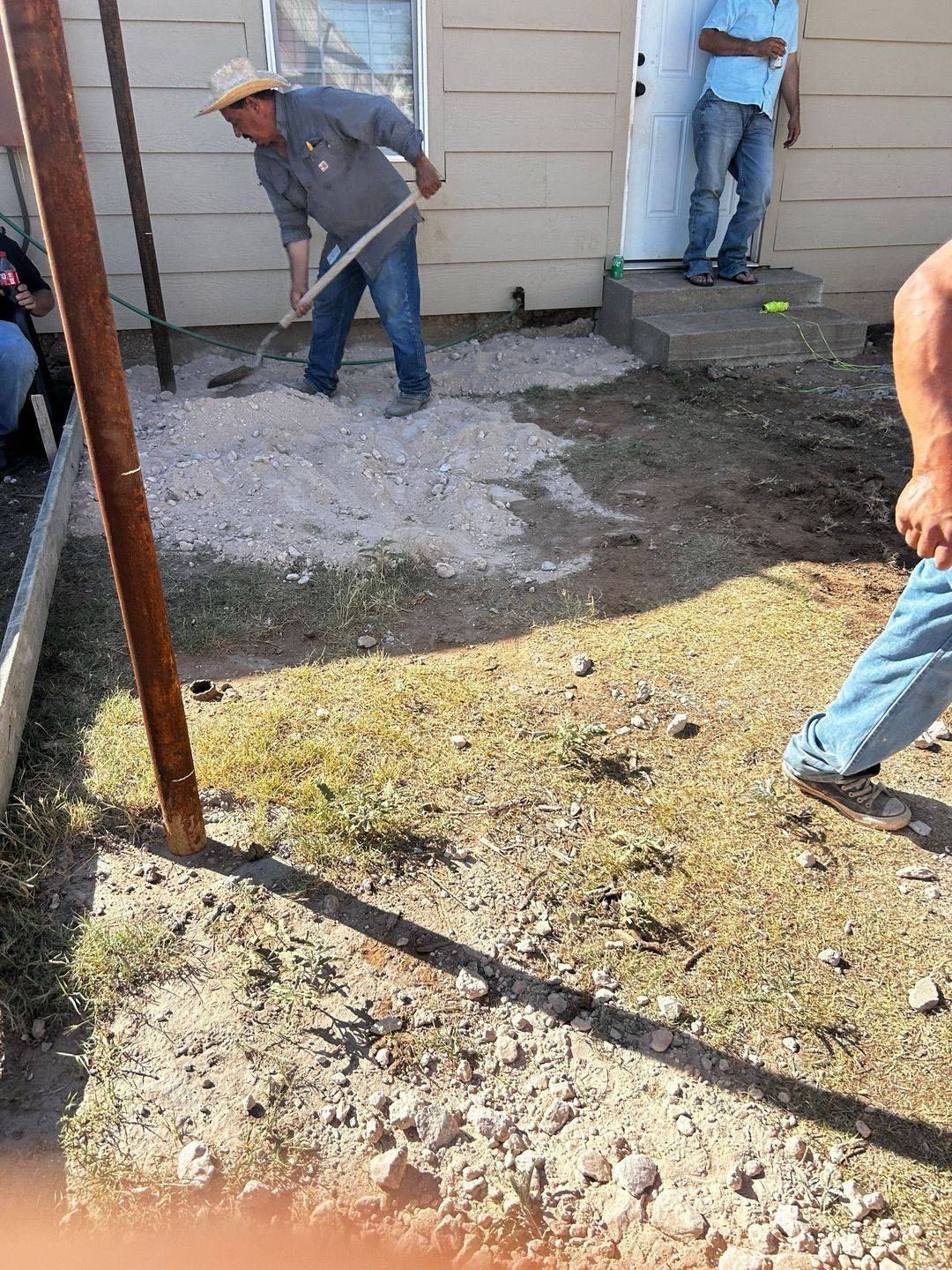 Men spreading gravel with rakes near a building entrance. One man wears a hat. Outdoors.