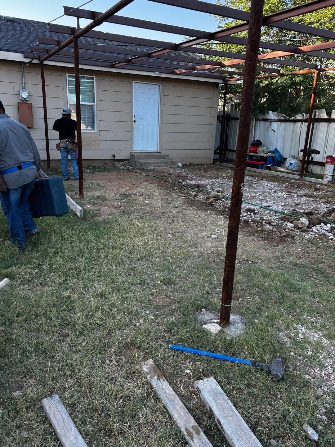 Workers near a small house with a metal pergola overhead; the ground is partly grass, partly dirt.