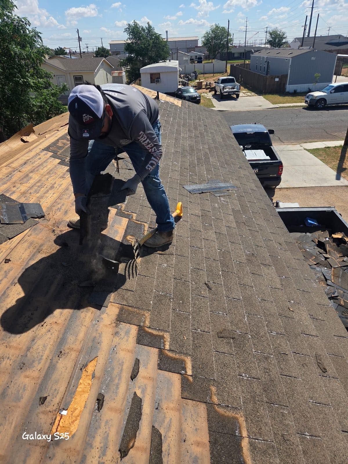 Roofer on a roof removing shingles, wearing a hat and jeans on a sunny day.