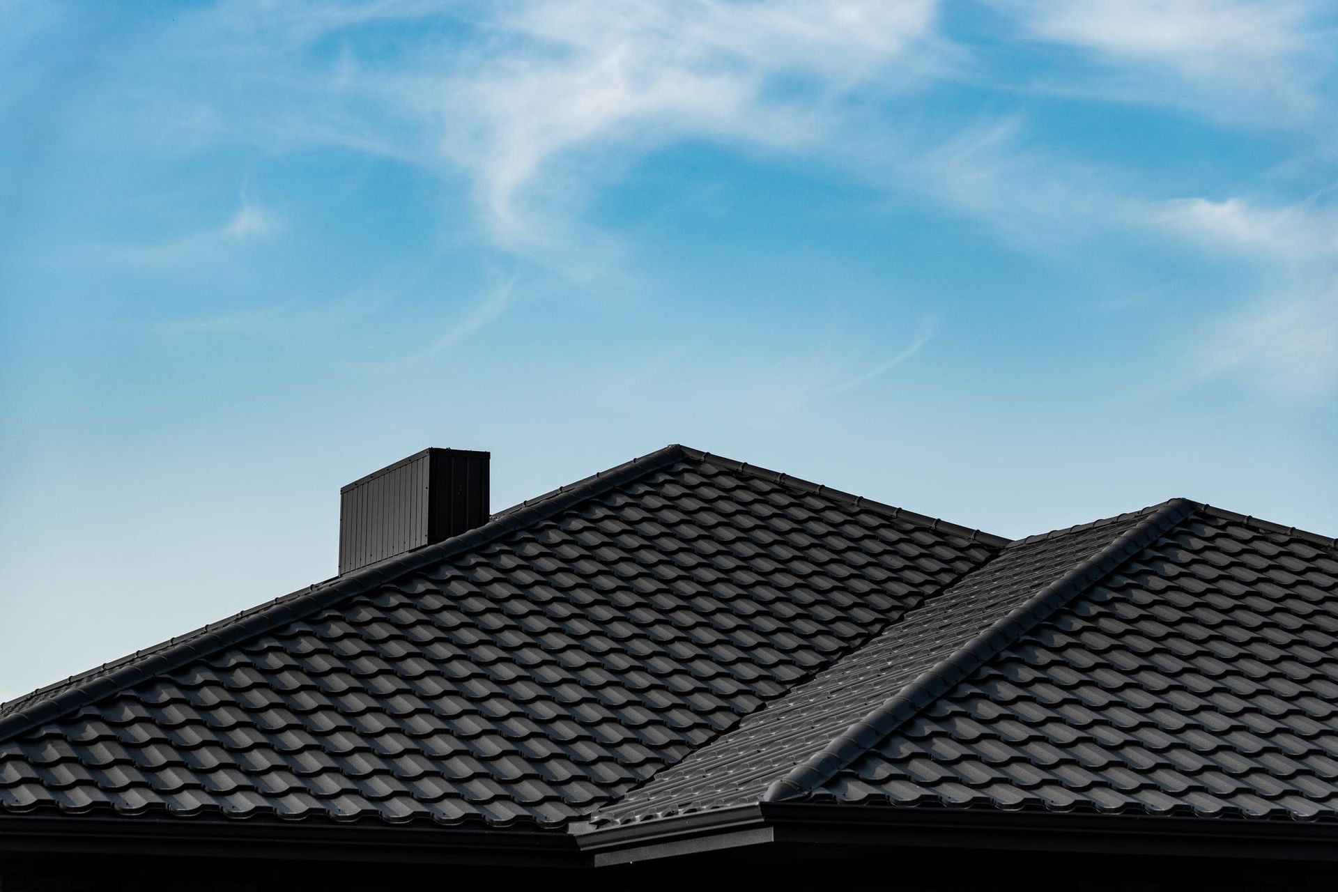 Black tiled roof with chimney against a bright blue sky with wispy clouds.
