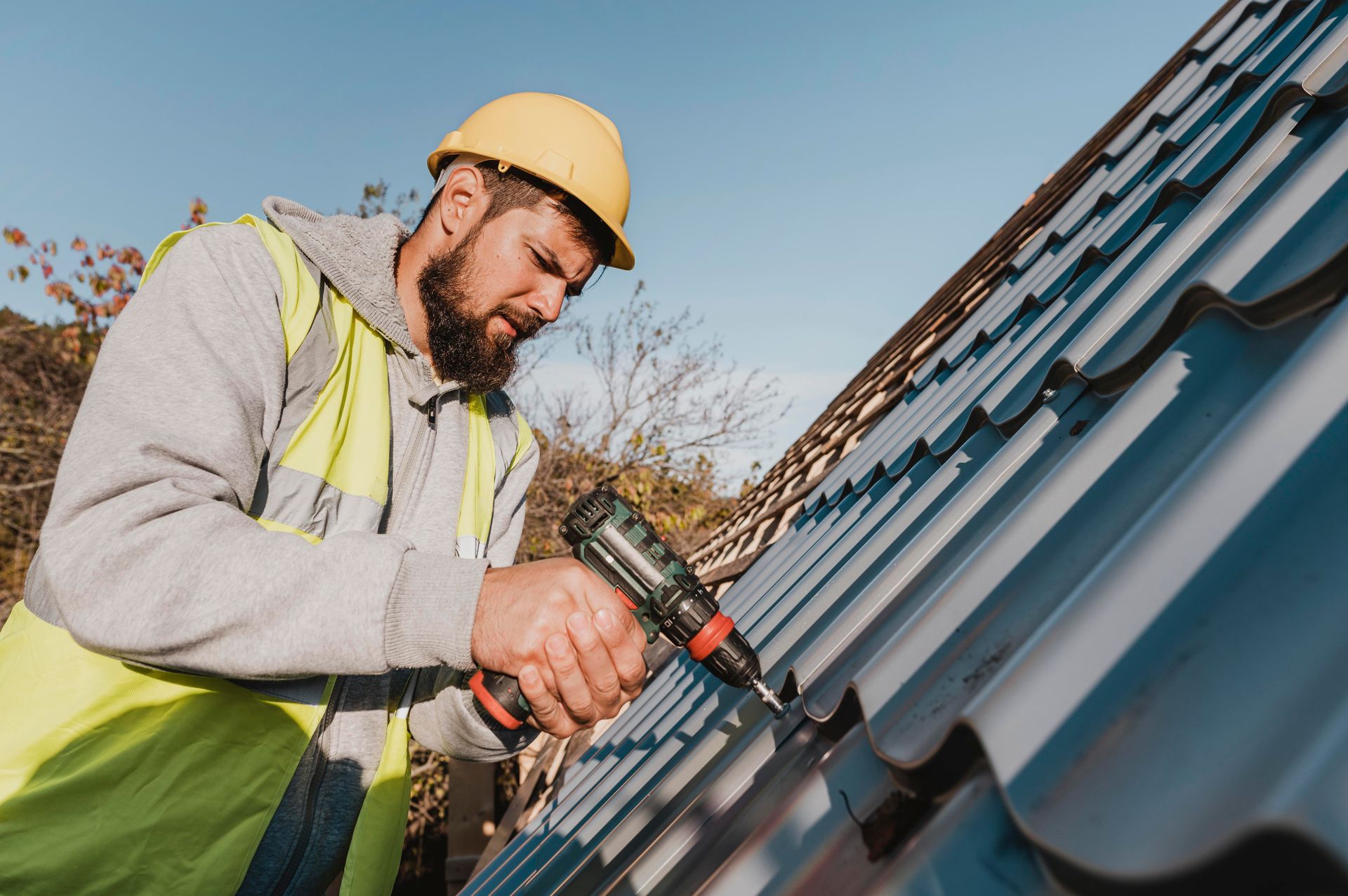 Construction worker in yellow hard hat and vest, installing metal roofing with a power drill.