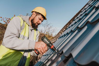 Construction worker in yellow hard hat and vest, installing metal roofing with a power drill.