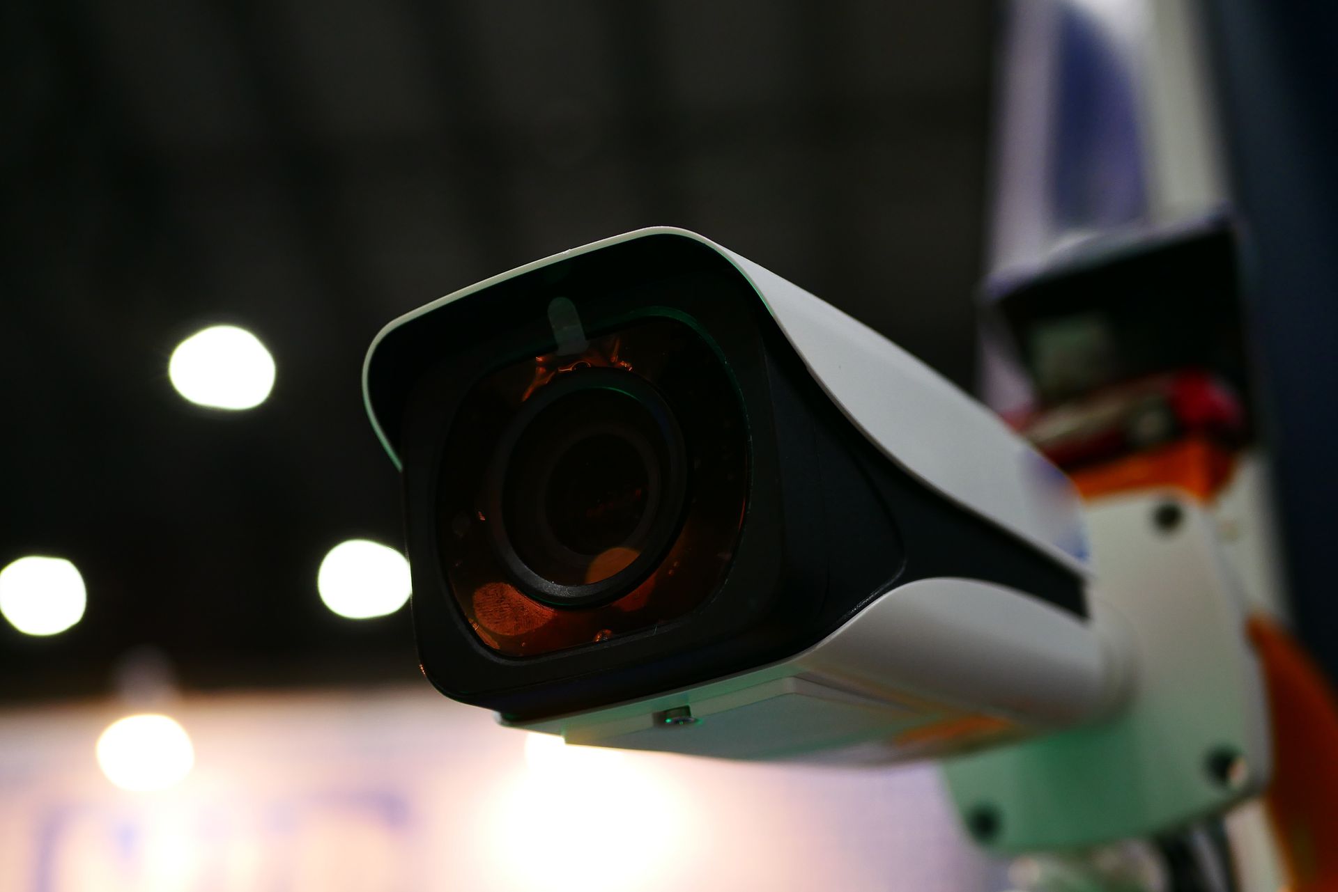 Close-up of a security camera with a black lens and white body, mounted on an orange and blue pole indoors.