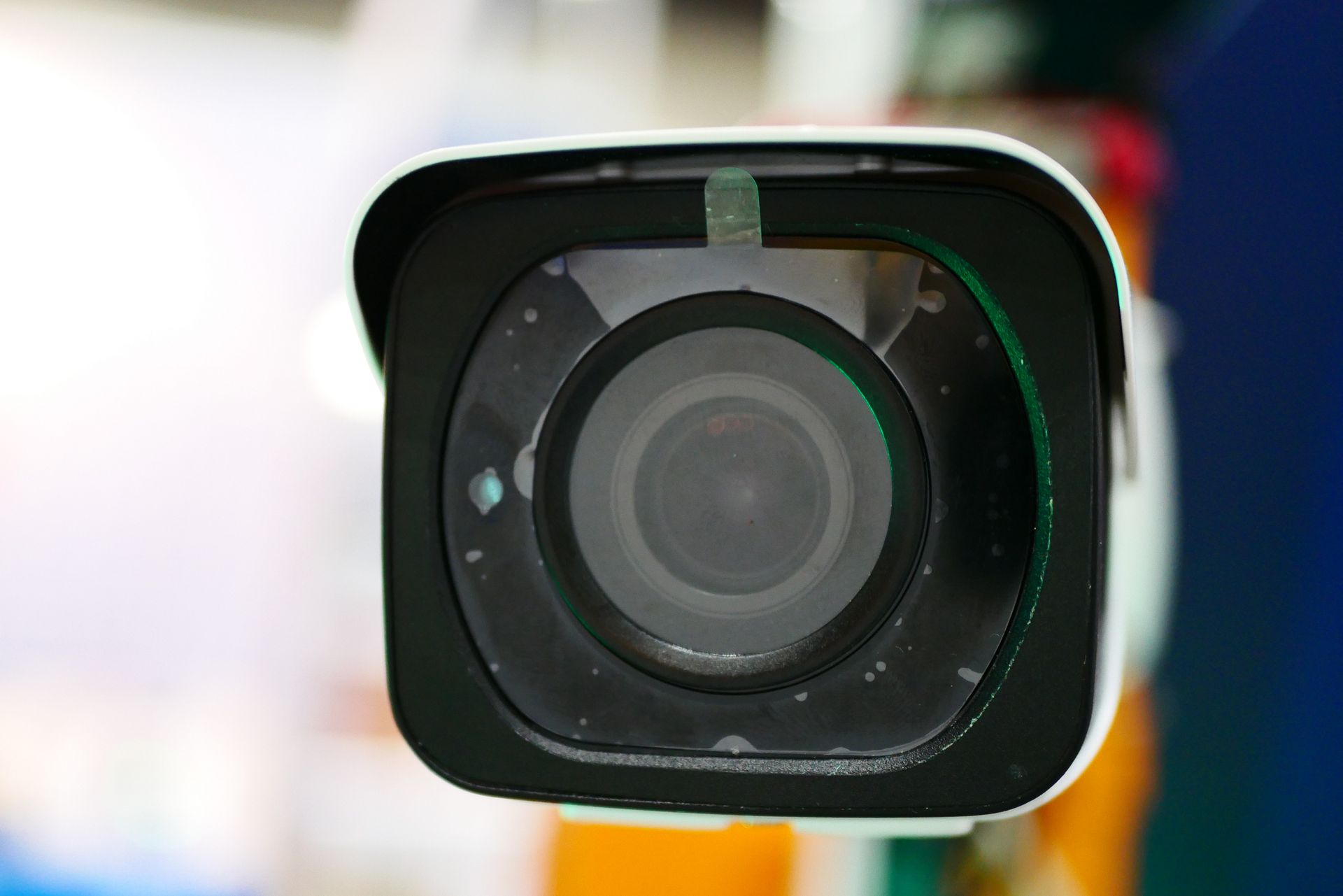 Close-up of a security camera with a black lens and white body, mounted on an orange and blue pole indoors.