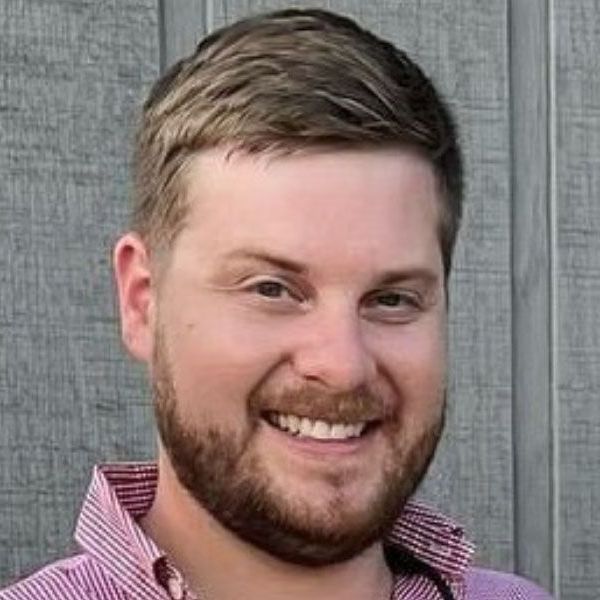 A man with a beard is smiling in front of a wooden wall.