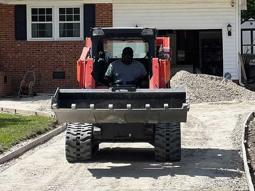 A man is driving a bulldozer down a dirt road in front of a house.