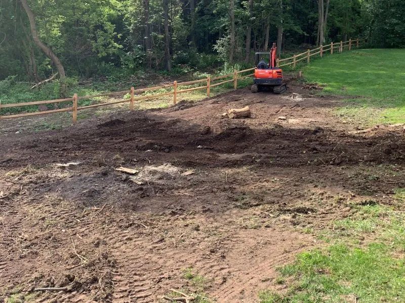 A red excavator is driving through a dirt field next to a wooden fence.