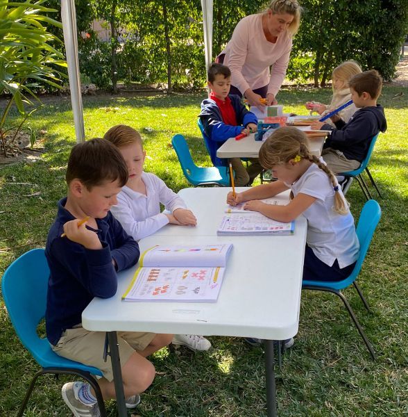 A group of children are sitting at tables outside in the grass.