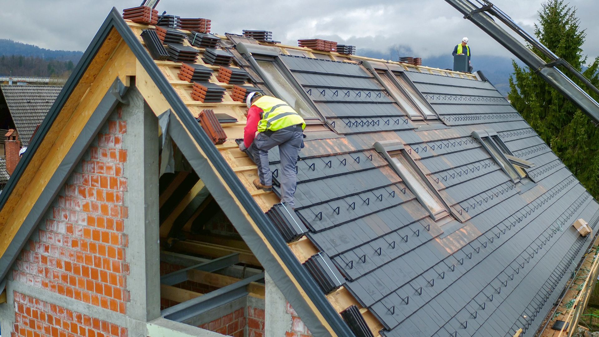Roofer installing dark gray tiles on a roof with skylights, wearing a safety vest. Brick wall and mountain backdrop.