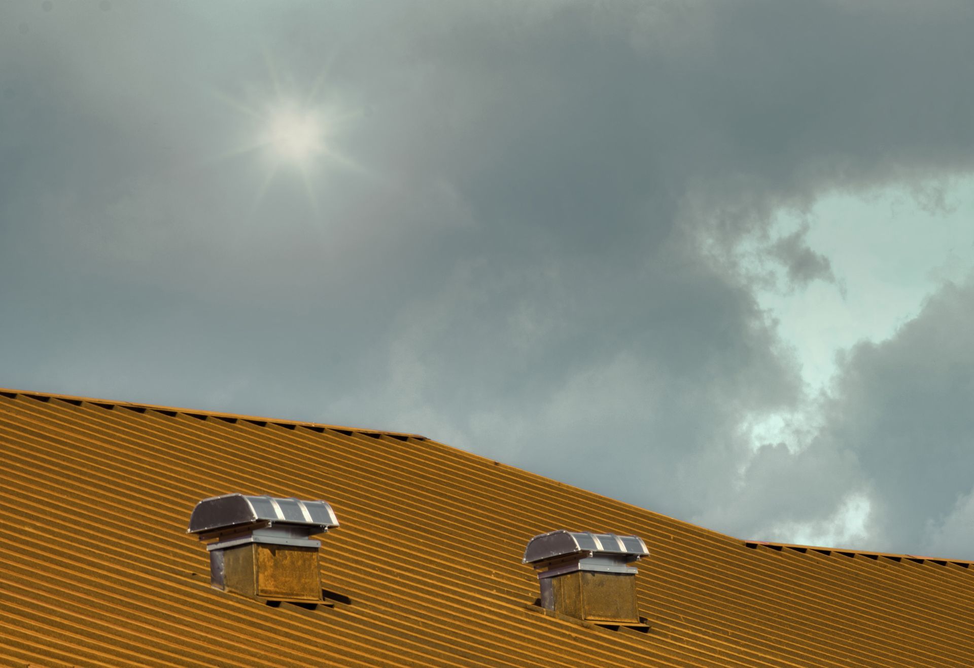 Yellow tiled roof with two chimneys against a cloudy sky, sun shining through.
