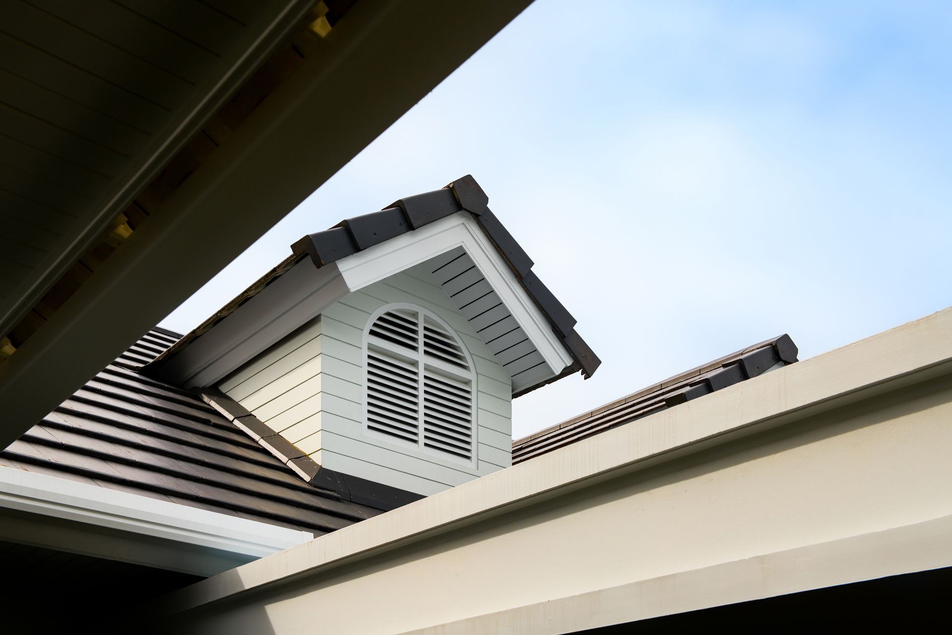 Dormer window with white siding and arched vent on a brown tile roof against a blue sky.