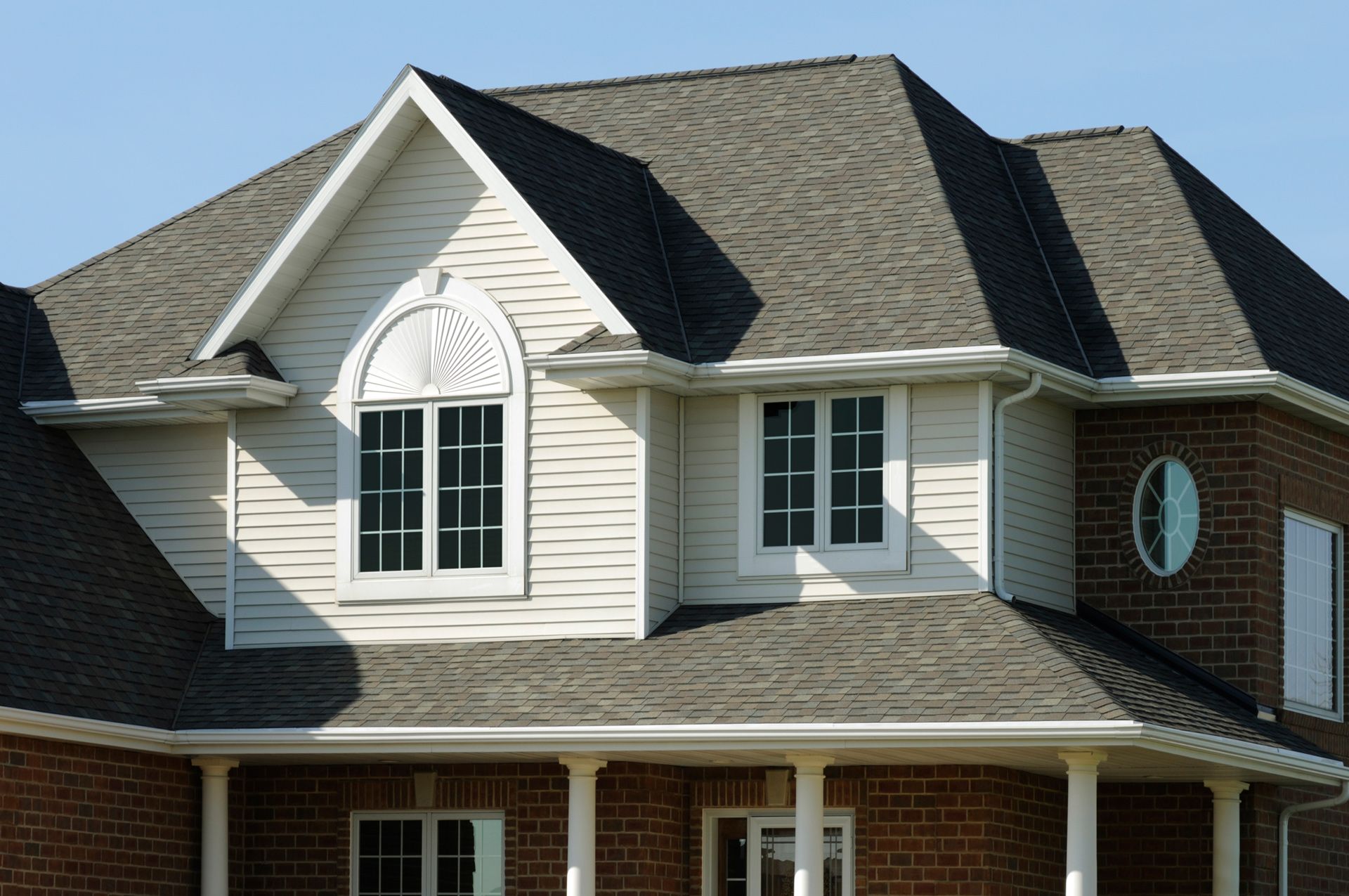 Two-story house with gray roof, white siding, and brick facade under a clear, blue sky.