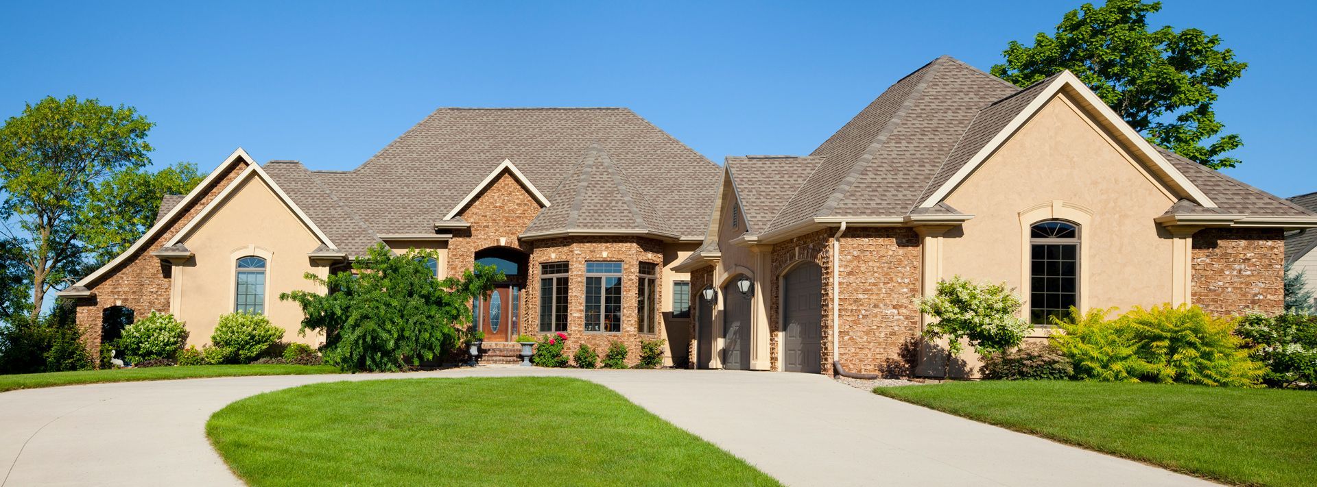 A large tan house with brick accents sits on a green lawn under a clear blue sky.
