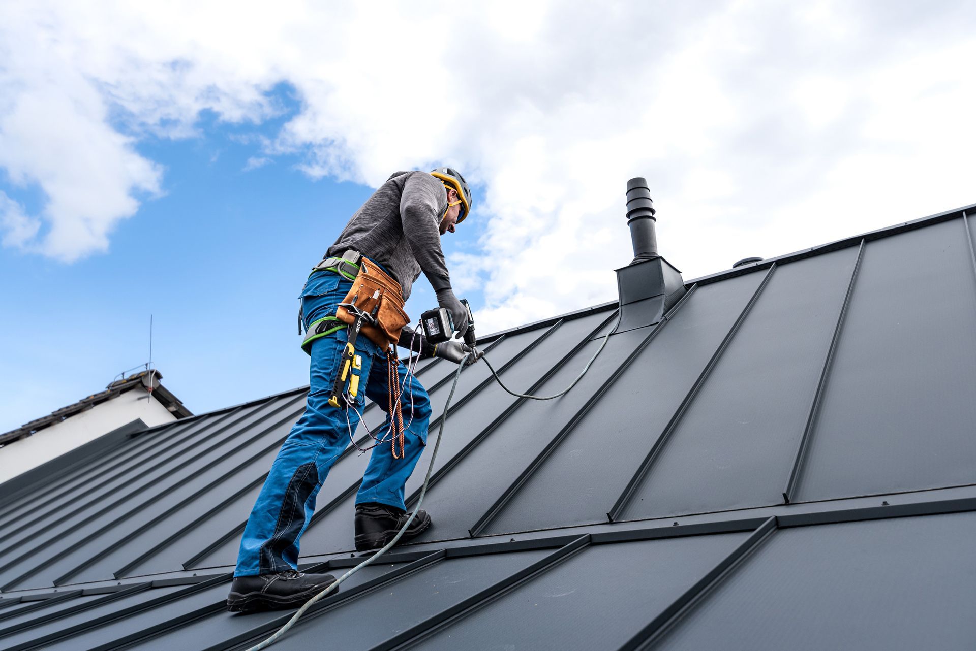 Roofer wearing safety gear working on a gray metal roof under a partly cloudy blue sky.