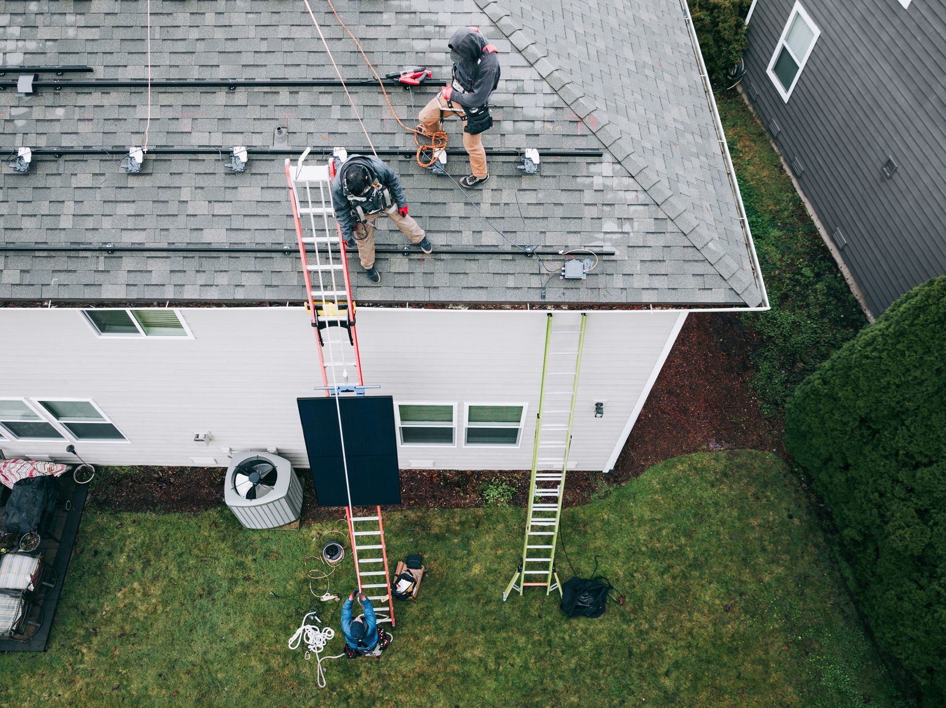 Two workers installing solar panels on a house roof, utilizing ladders and safety harnesses, green grass below.