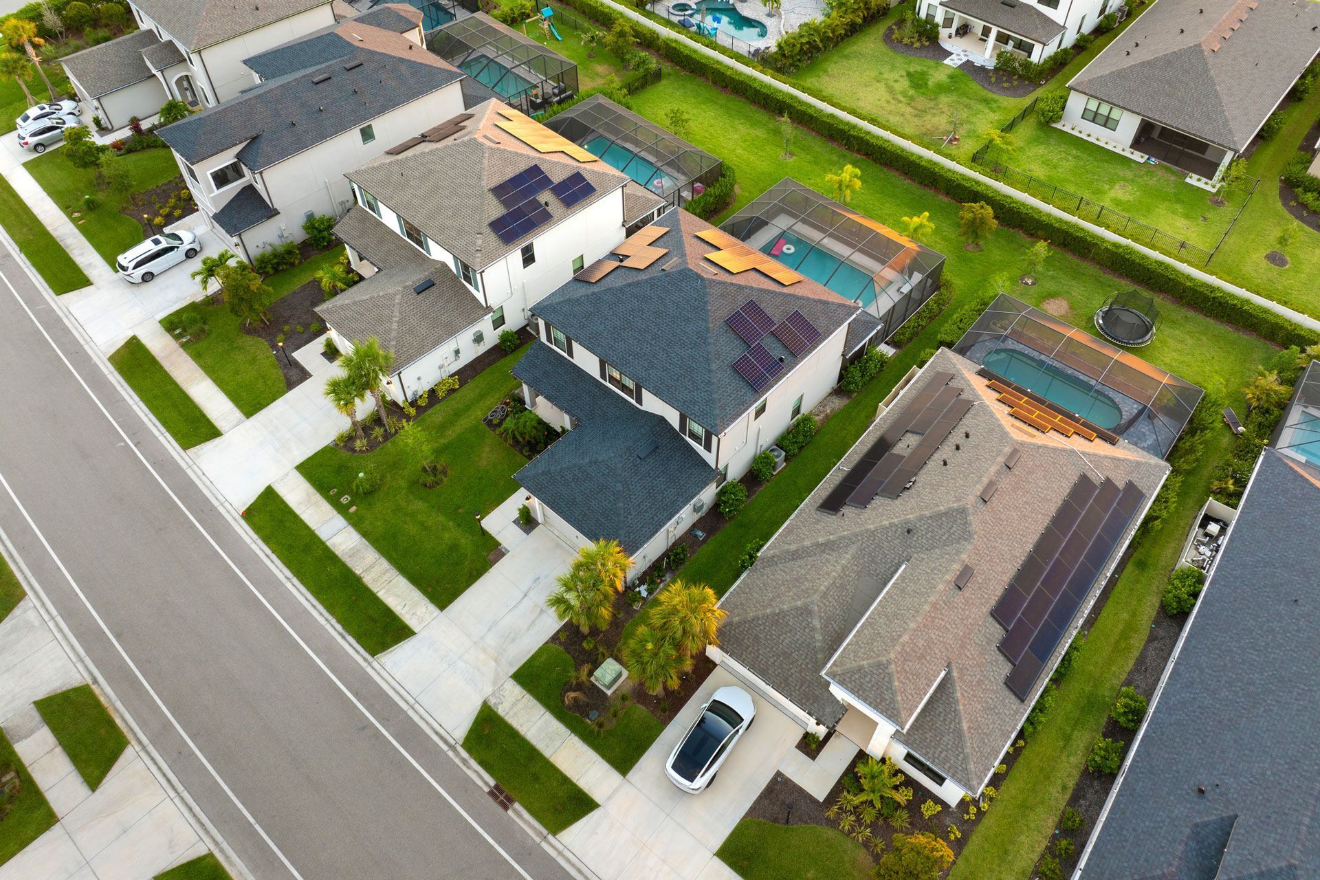 Aerial view of houses with solar panels, pools, and driveways along a paved street.