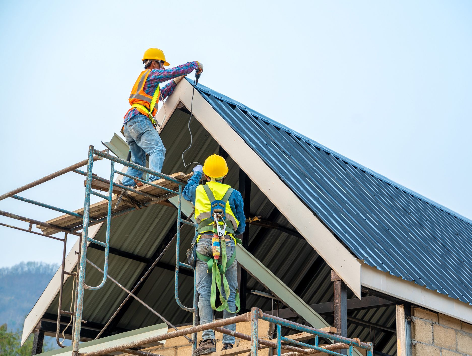 Two construction workers on scaffolding installing a metal roof, wearing safety gear and yellow hard hats.