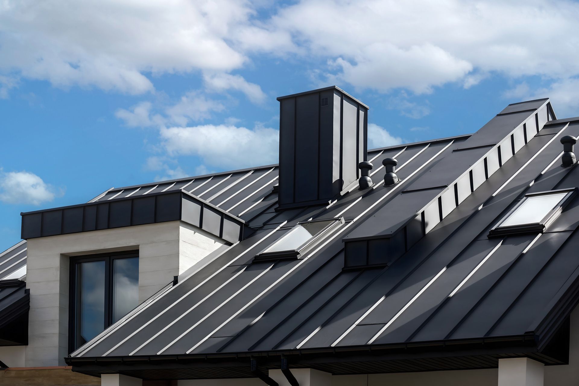 Dark metal roof with chimney and skylights against a cloudy blue sky.