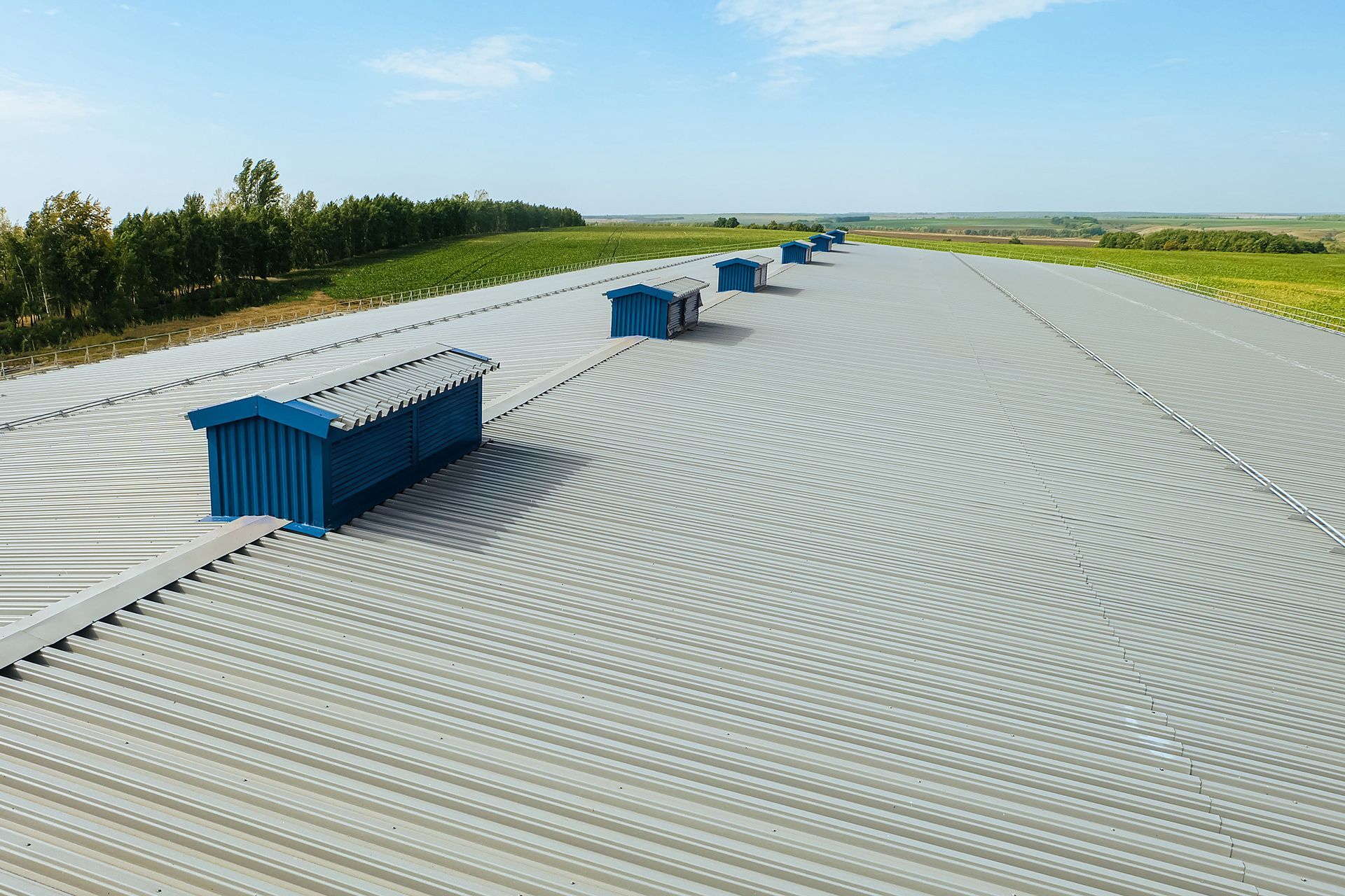 A long, gray corrugated roof with blue storage sheds and fields in the background under a blue sky.