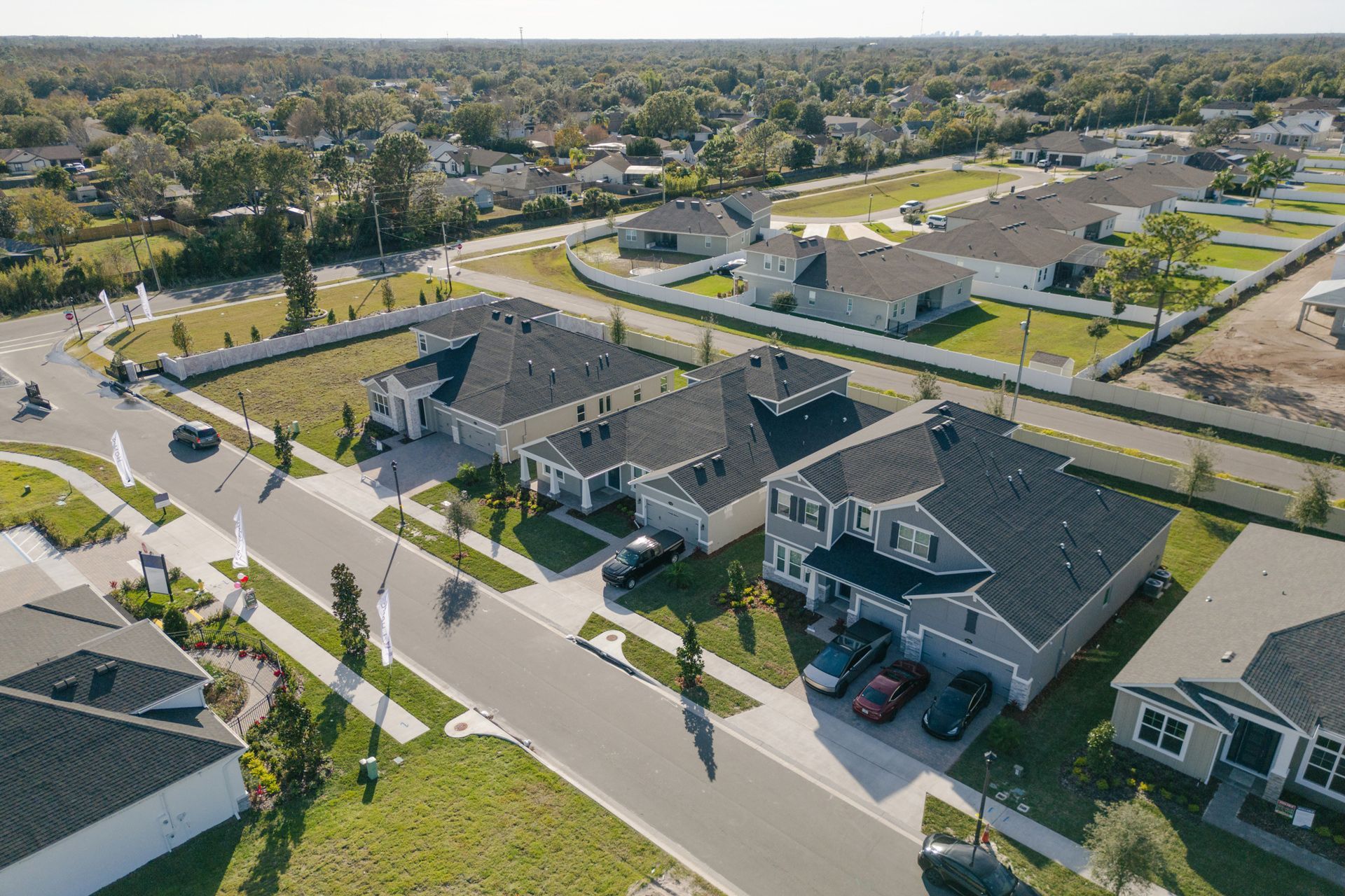 Aerial view of a suburban neighborhood with houses, trees, roads, and cars under a blue sky.
