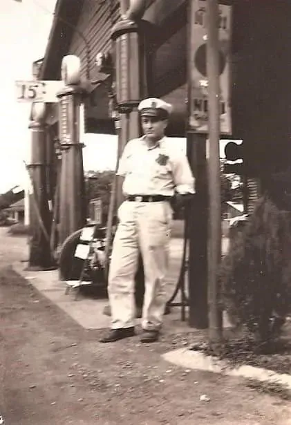 A black and white photo of a man standing next to a gas pump.