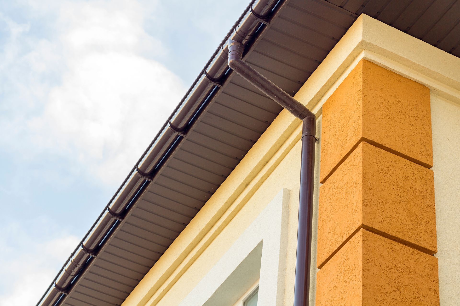 Corner of a house with beige siding, white trim, and a dark roof gutter against a cloudy sky
