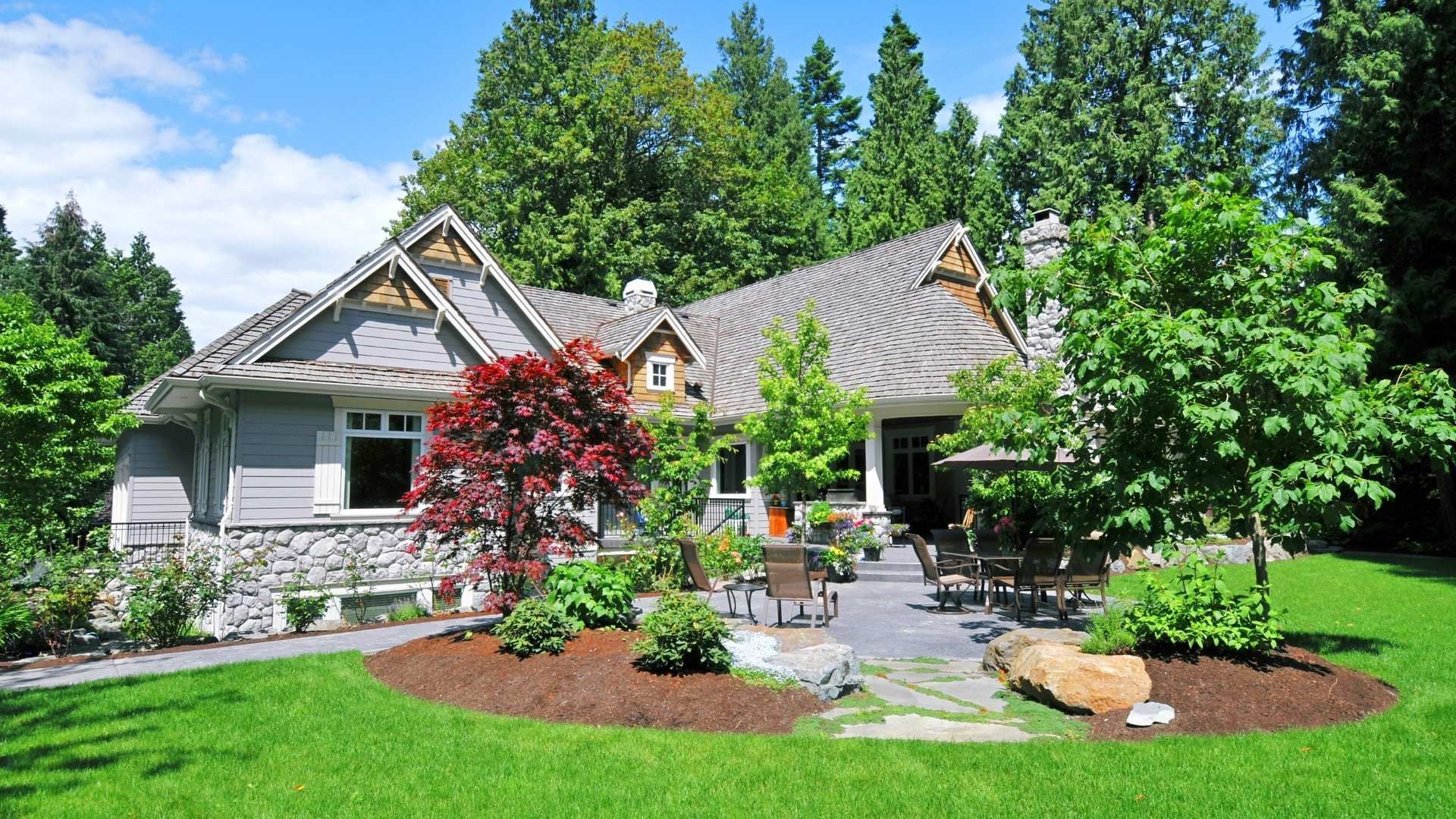 Gray house with a stone and wood facade, patio, and lush green landscaping under a blue sky.