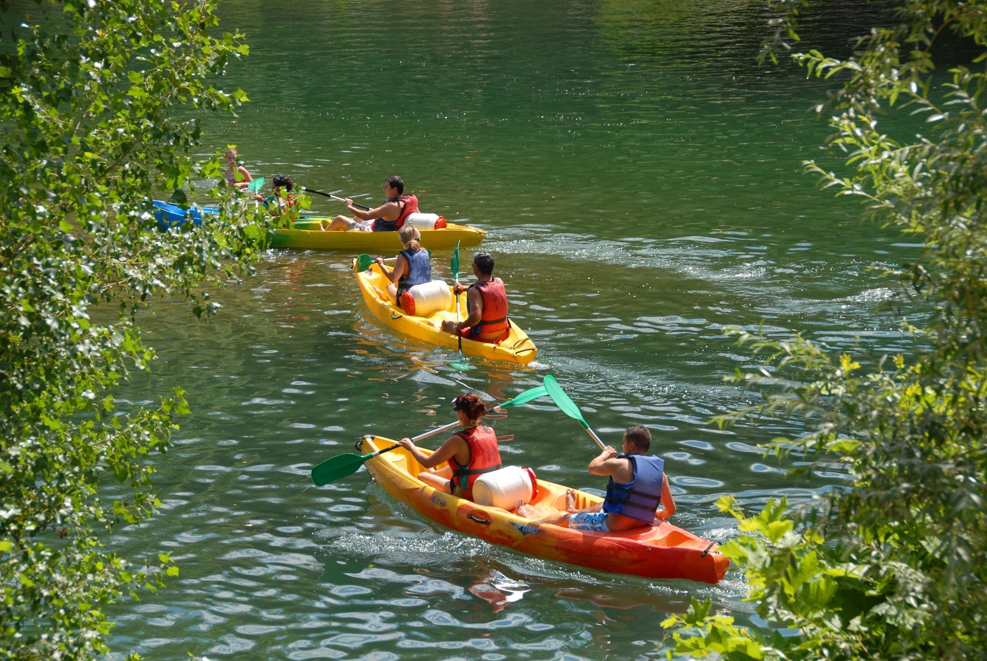 People paddling colorful canoes on a calm green river in a wooded area