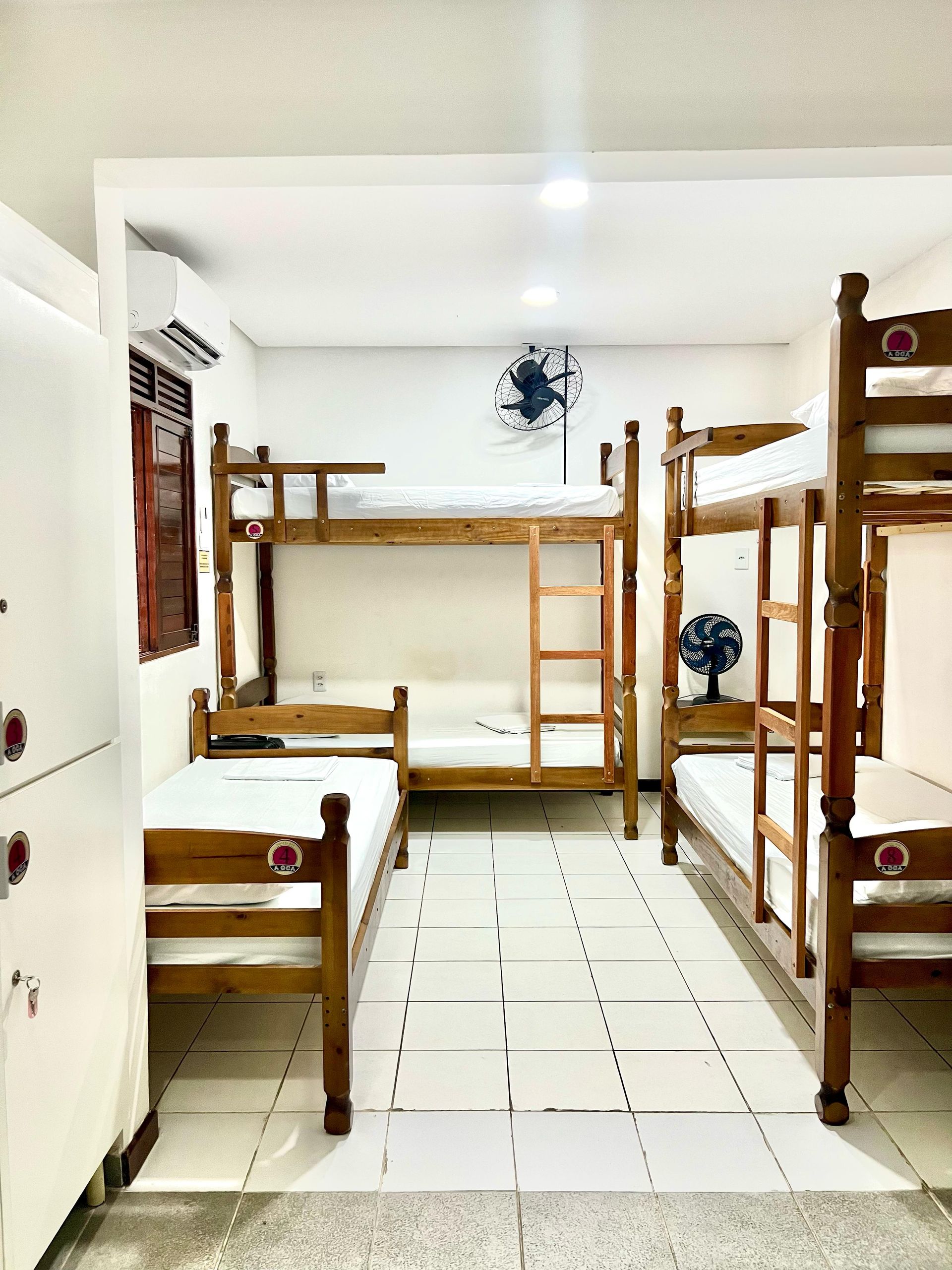 Quarto de dormitório com duas camas de beliche de madeira, uma escrivaninha, uma escada e piso de azulejos brancos.