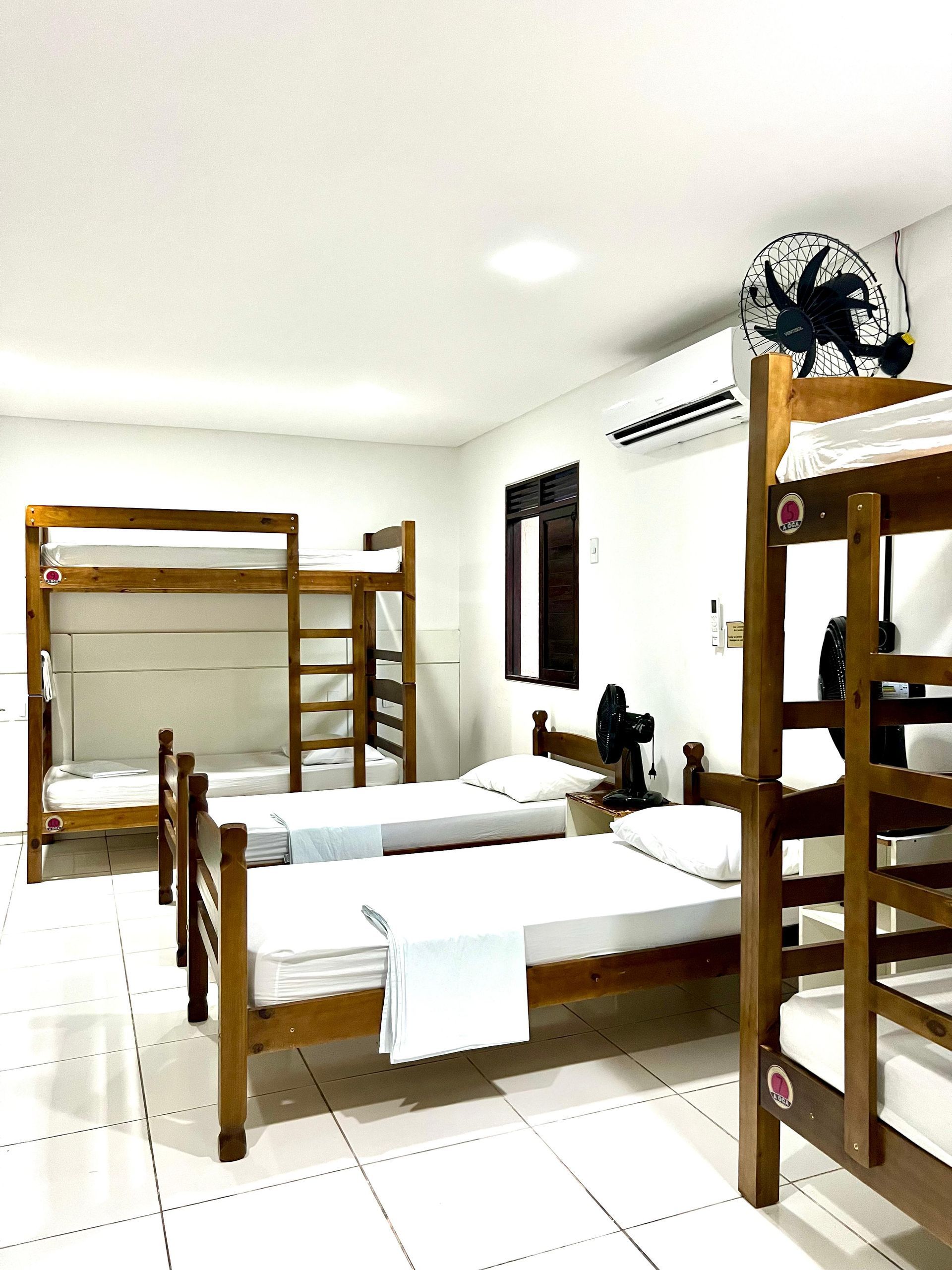 Quarto de dormitório com beliches de madeira, roupa de cama branca, piso de azulejo e ventilador de teto.