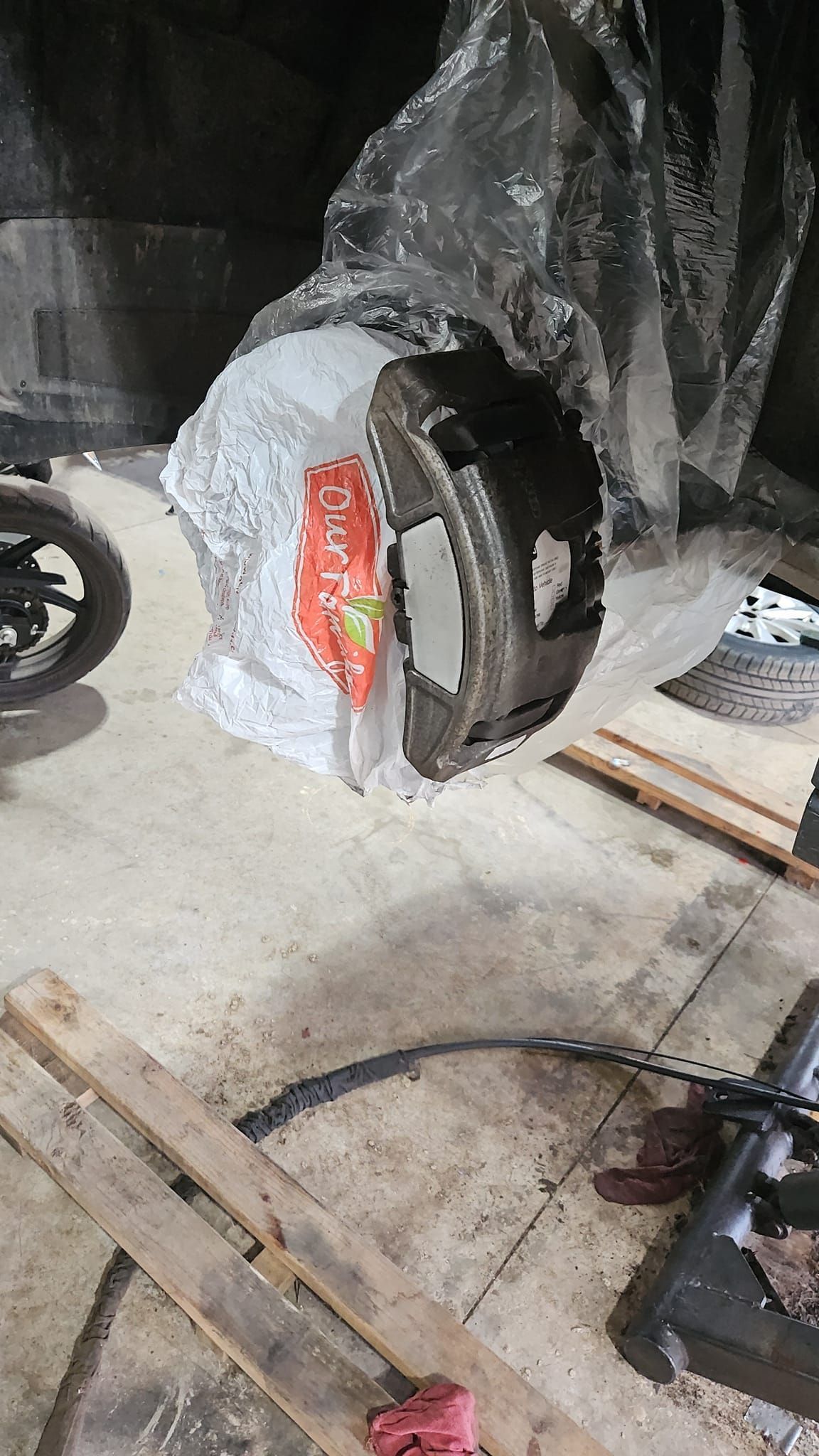 A wrapped car tire on a concrete floor next to wooden planks, a motorcycle wheel, and tools.