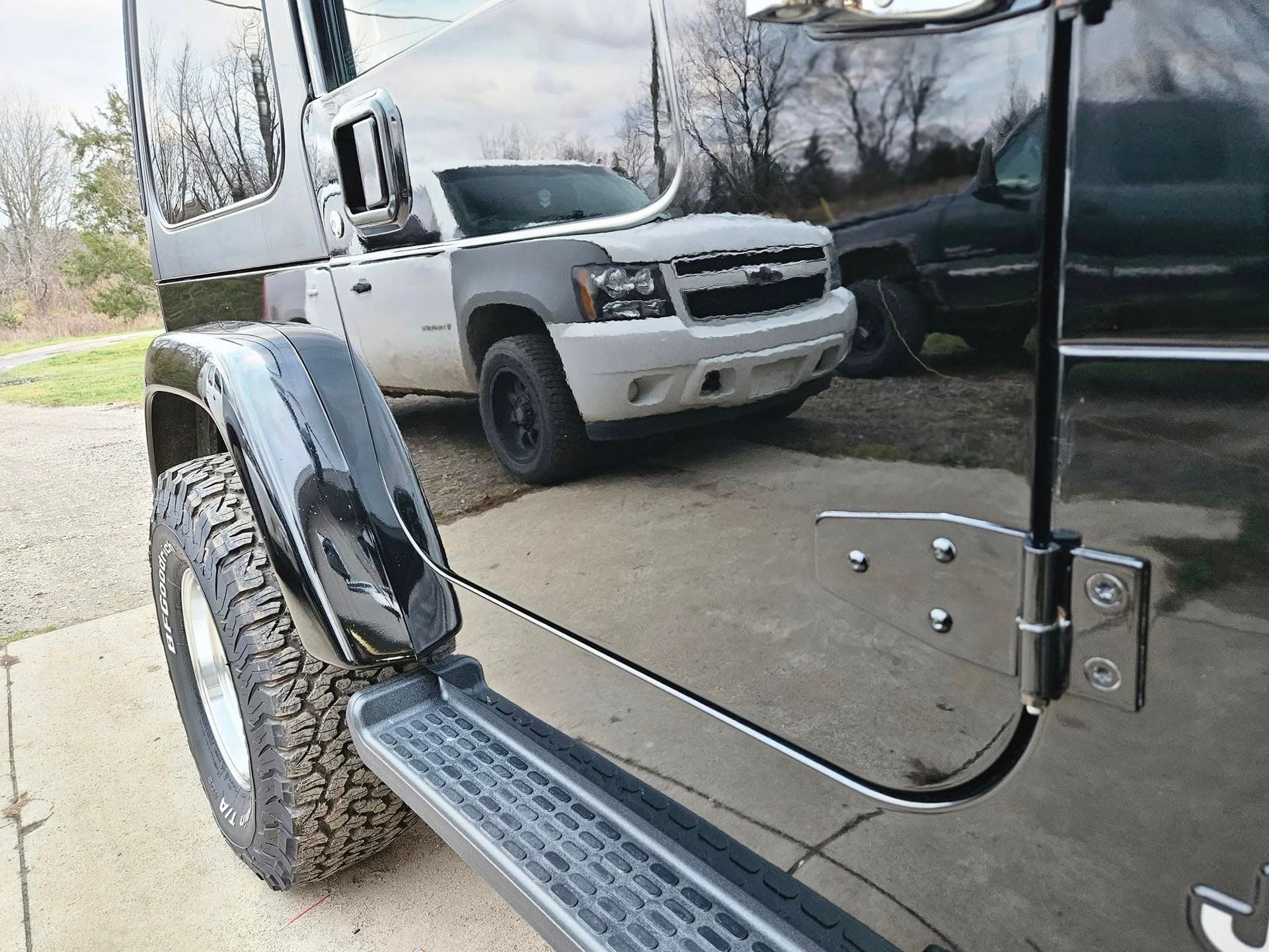 Black Jeep's side and tire with a white SUV in the background, reflected on the Jeep's surface.