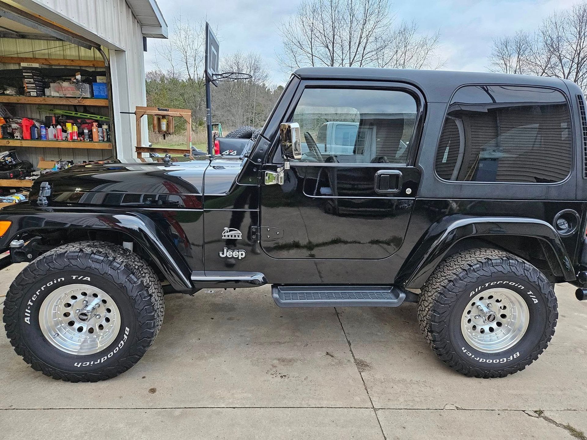 Black Jeep Wrangler with hardtop and chrome wheels parked in front of a garage.