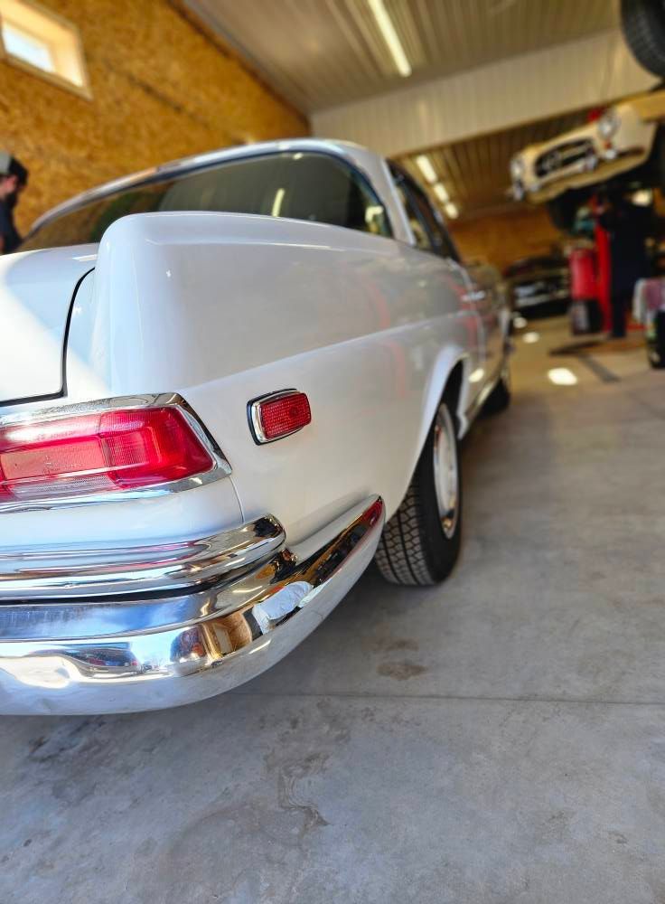 White classic car's rear quarter panel in a garage. Chrome bumper, red taillight, concrete floor, overhead lights.