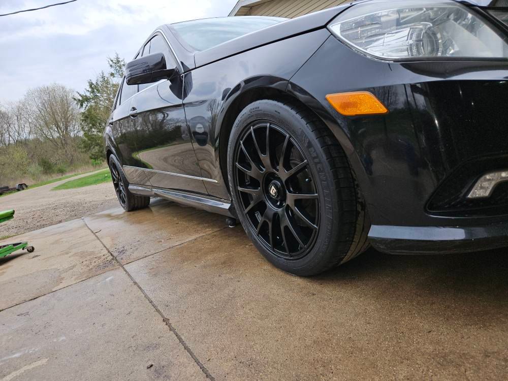 Black car parked on a concrete surface; black wheels, orange side marker, cloudy day.