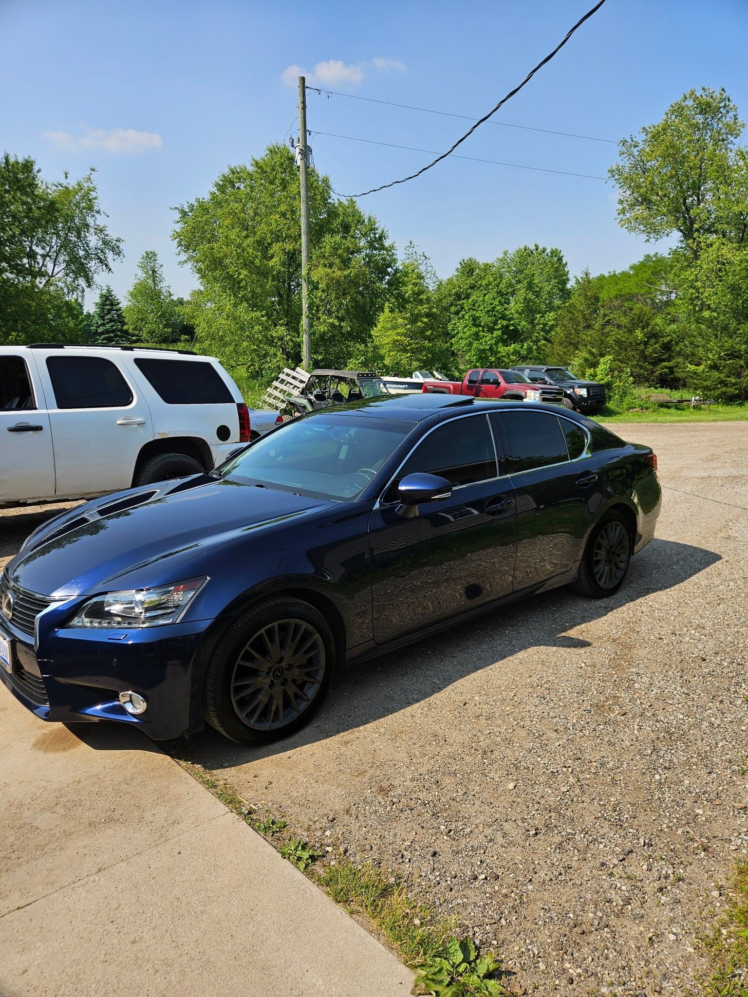 Blue Lexus sedan parked on gravel in front of trees and a white SUV on a sunny day.