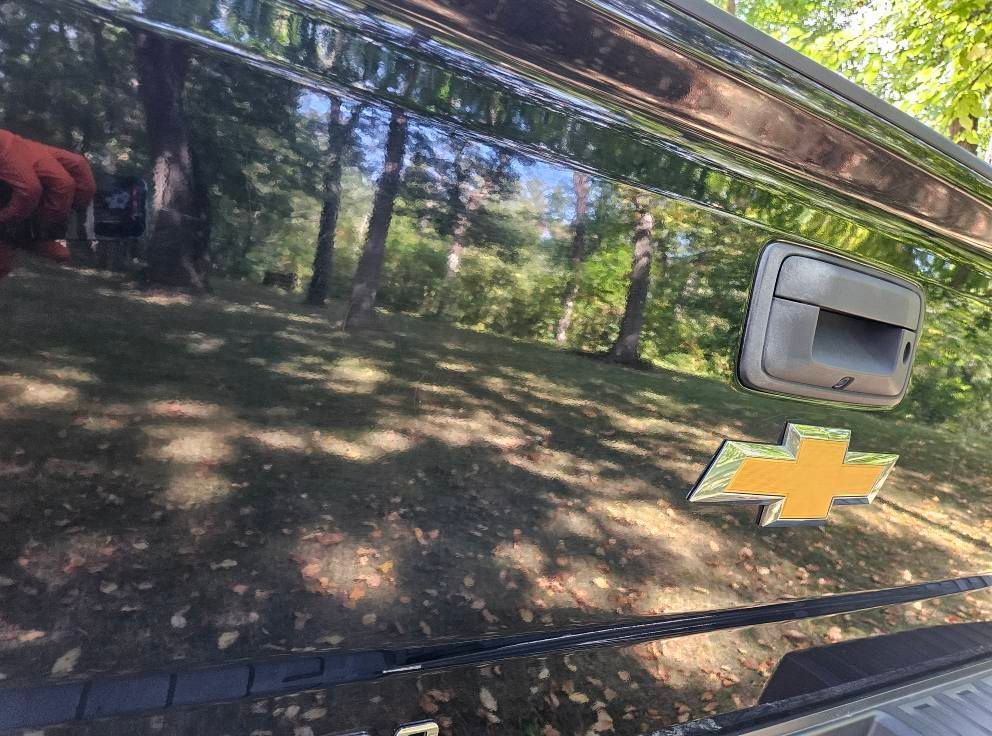 Black Chevrolet truck bed with handle, gold emblem, reflecting trees and grass.