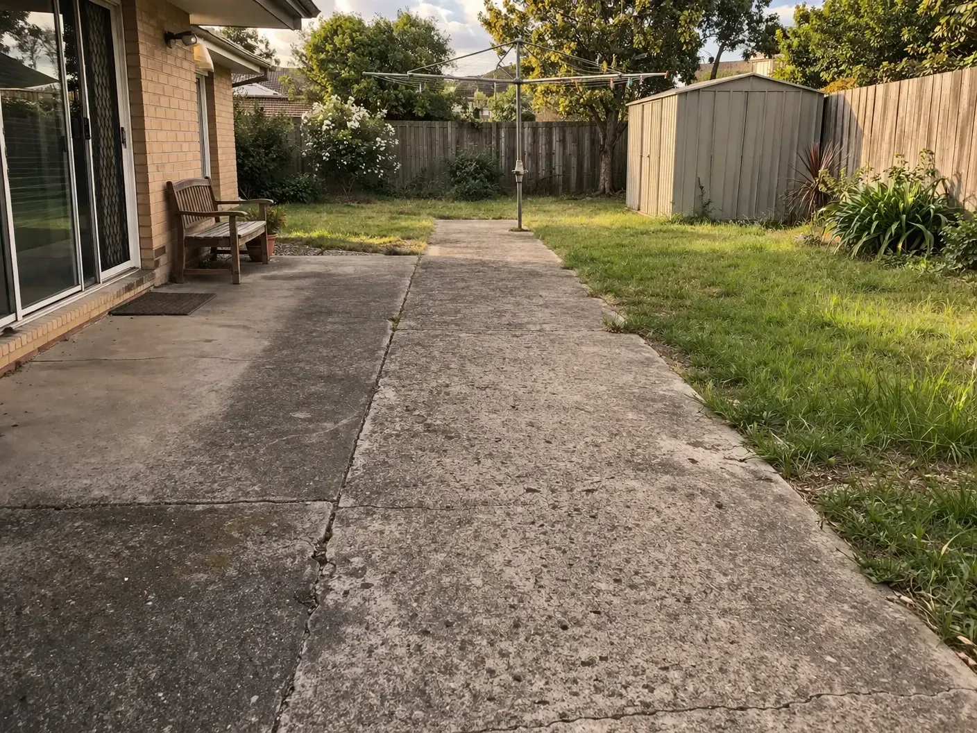 A worn concrete patio and walkway lead through a grassy backyard toward a shed, with a wooden bench near Drayton, Toowoomba