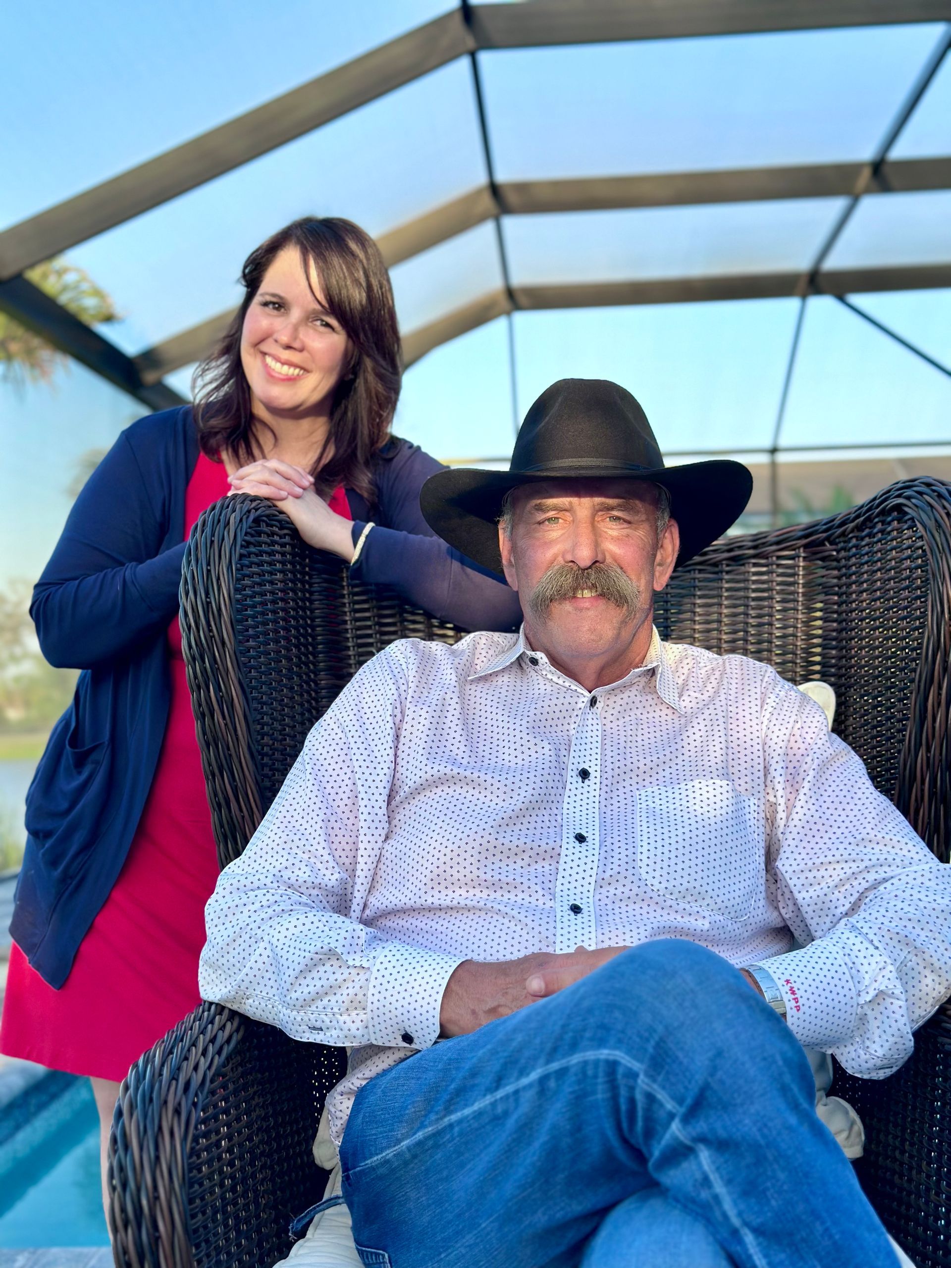 Woman in red dress and cardigan leans on a man in a cowboy hat and jeans, seated in a wicker chair outdoors.
