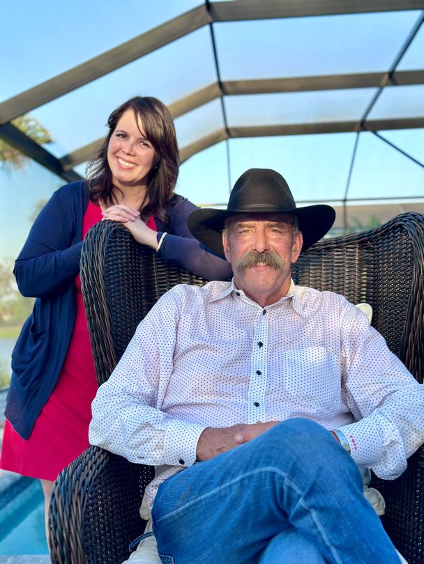 Woman in red dress and cardigan leans on a man in a cowboy hat and jeans, seated in a wicker chair outdoors.