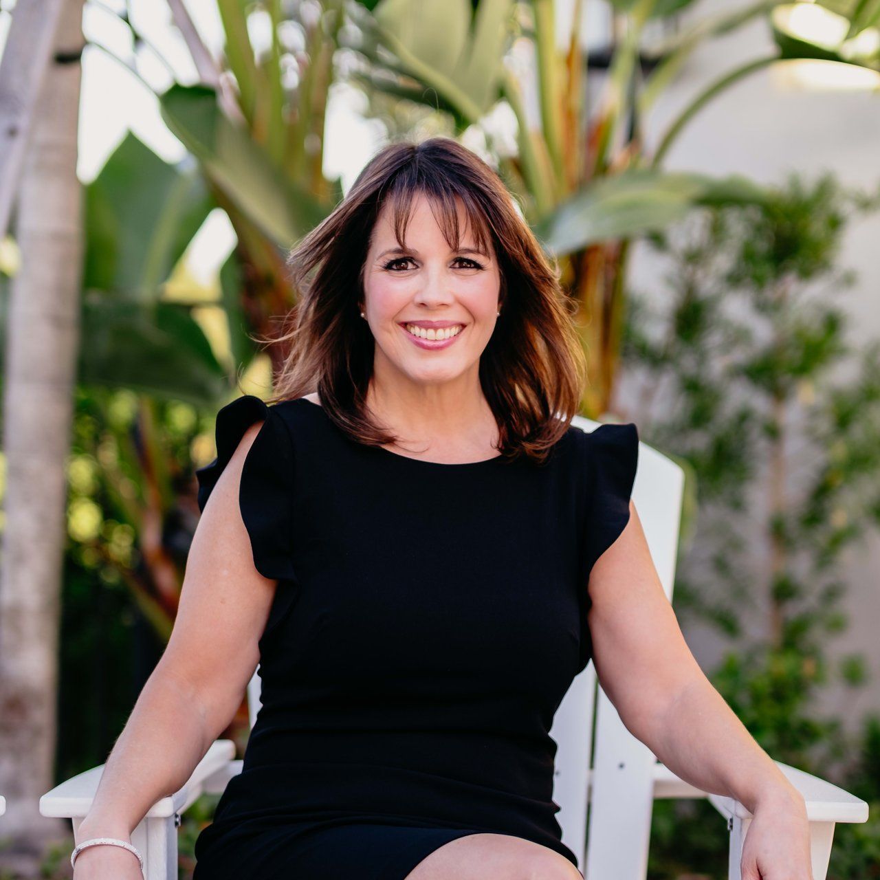 Woman with brown hair and a black dress smiles while sitting outside.
