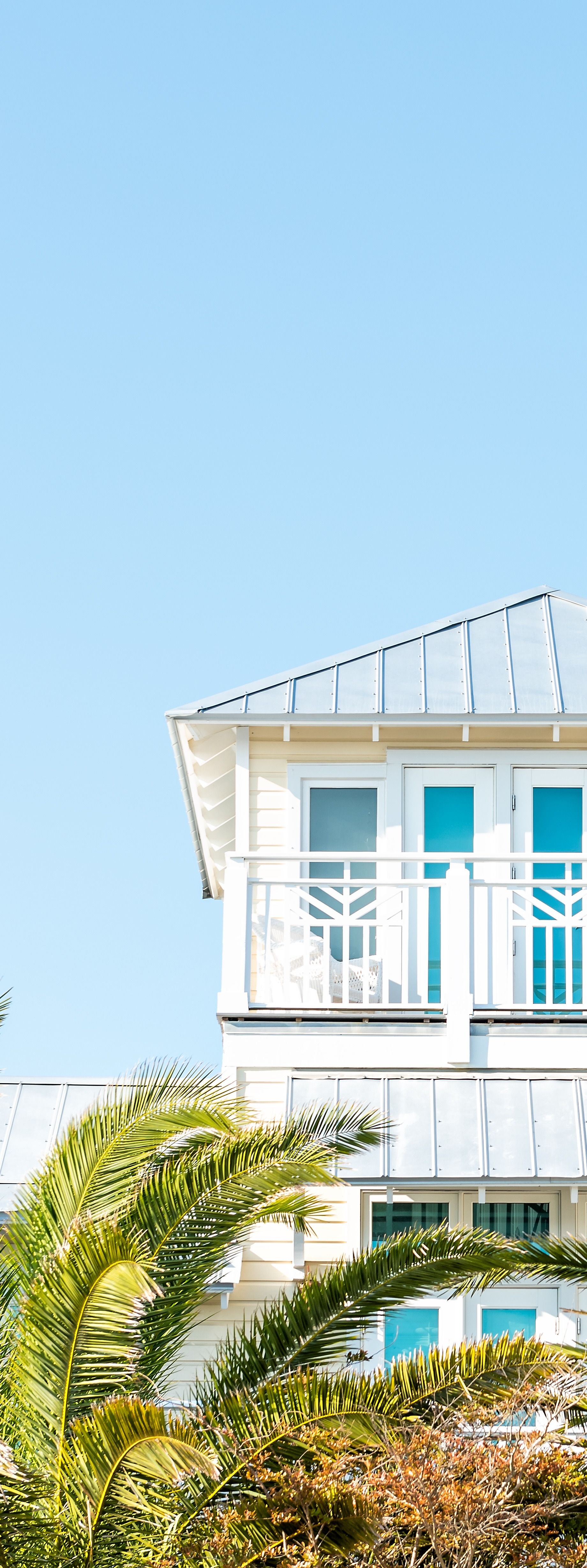 White beach house with blue windows against a clear blue sky. Green foliage in the foreground.