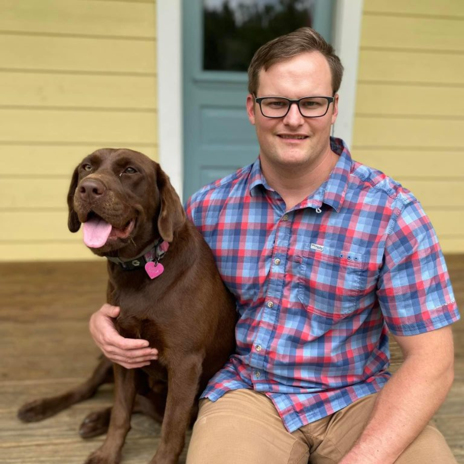 Dr. Fold in plaid shirt and glasses sits with brown Labrador on porch.