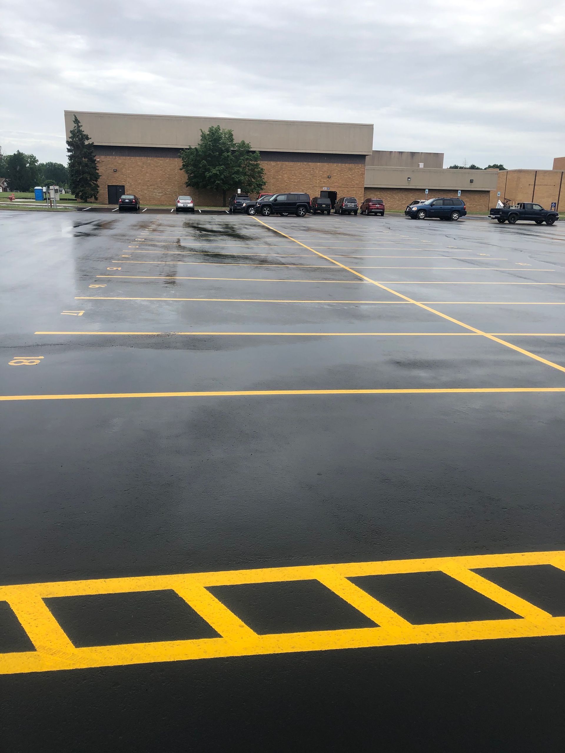 Wet parking lot with yellow lines. Cars are parked near a brick building under a cloudy sky.