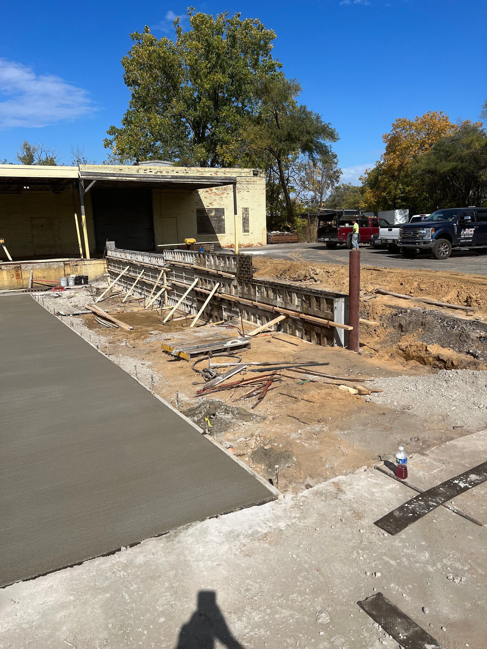 Construction site with concrete forms next to a new concrete slab, with a building and parked vehicles in the background.