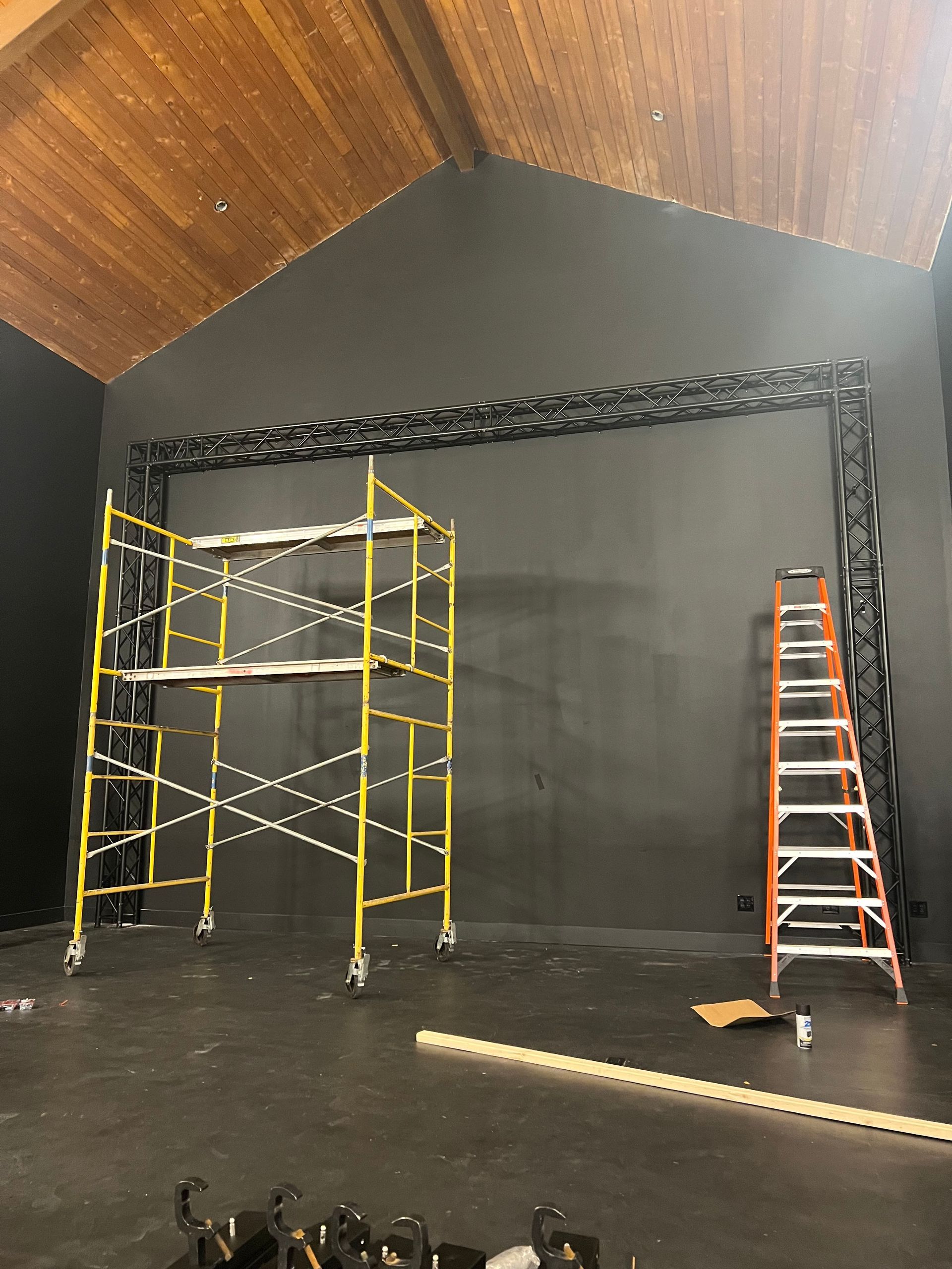 A stage with scaffolding, a ladder, and black wall with trussing. Wooden ceiling.