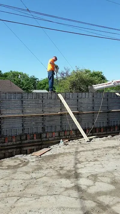 Construction site: a worker trowels concrete in a long trench reinforced with metal rods, along a wall and a sidewalk.